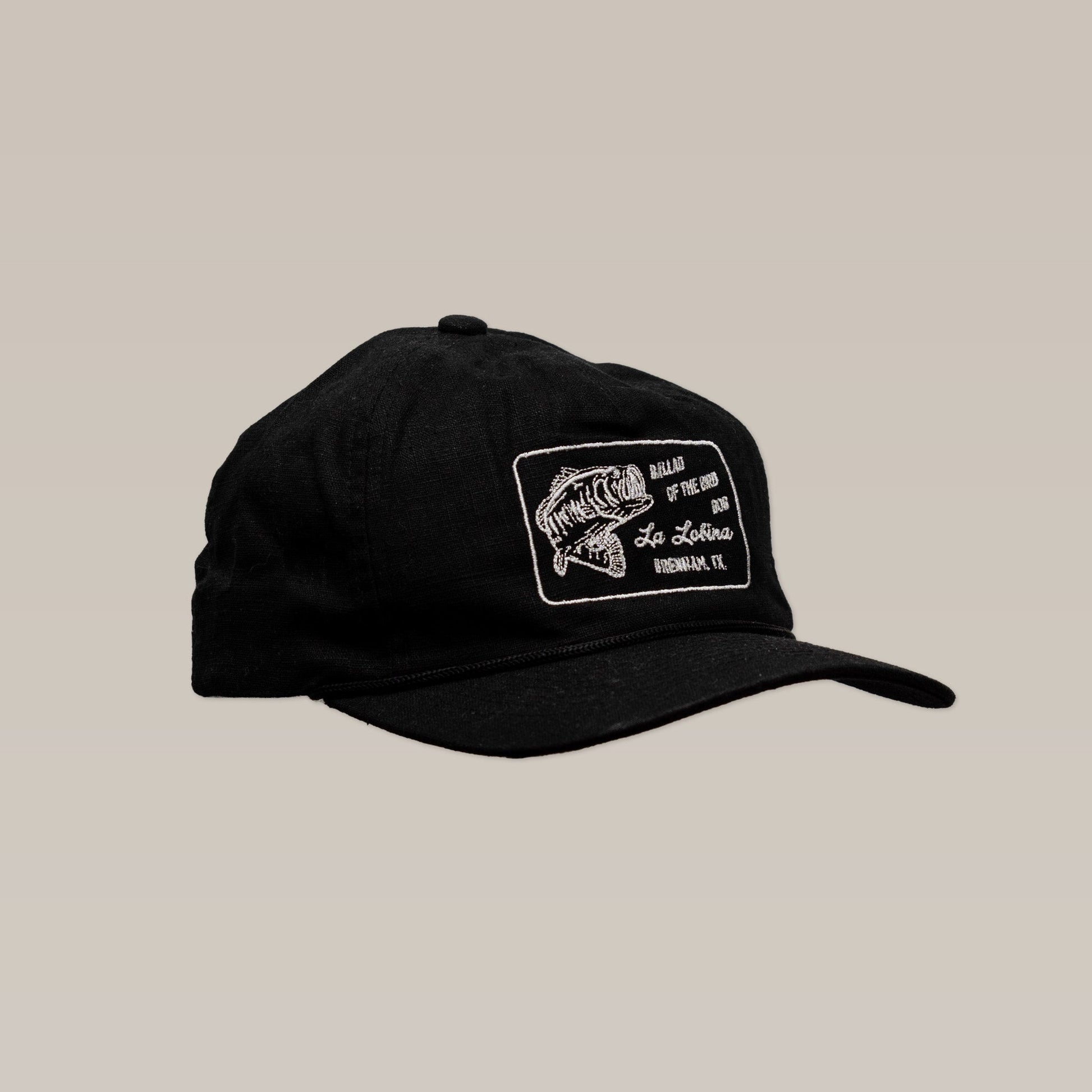 The Ballad of the Bird Dog La Lobina Shop Hat is a black fishing cap featuring a white embroidered fish logo and text patch on the front, shown against a neutral background.