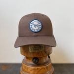 The Shop Hat | Merit Badge by Ballad of the Bird Dog is a brown snapback cap with a dog merit badge patch, shown on a vintage wooden hat stand.