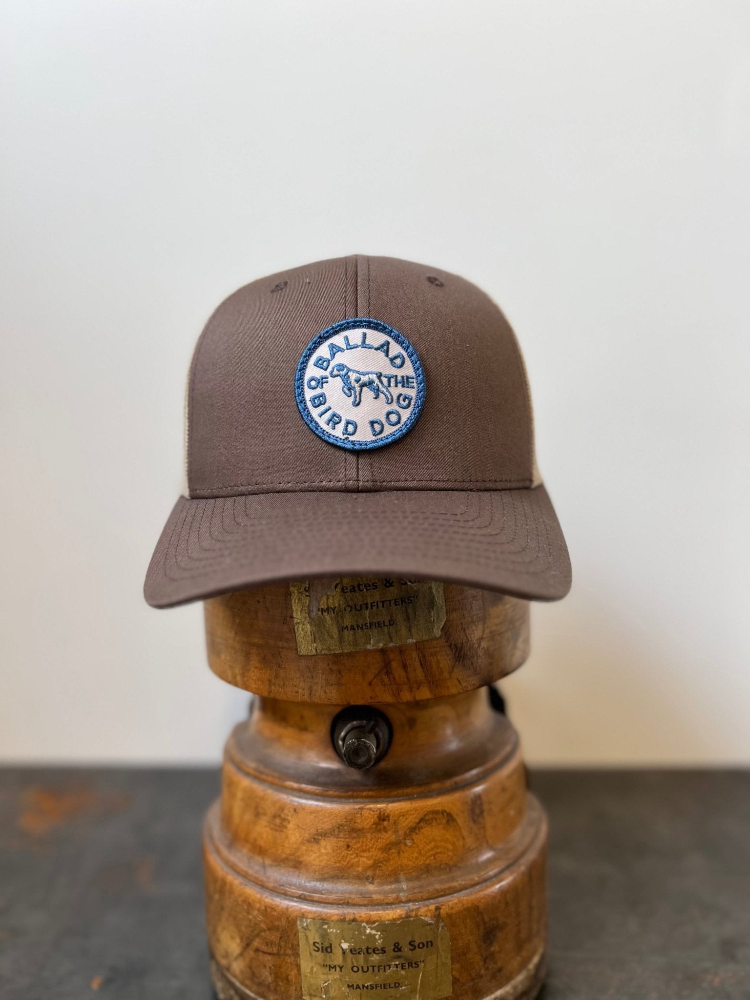 The Shop Hat | Merit Badge by Ballad of the Bird Dog is a brown snapback baseball cap featuring a circular blue and white "Ballad of the Bird Dog" patch on the front, shown on a wooden hat stand.