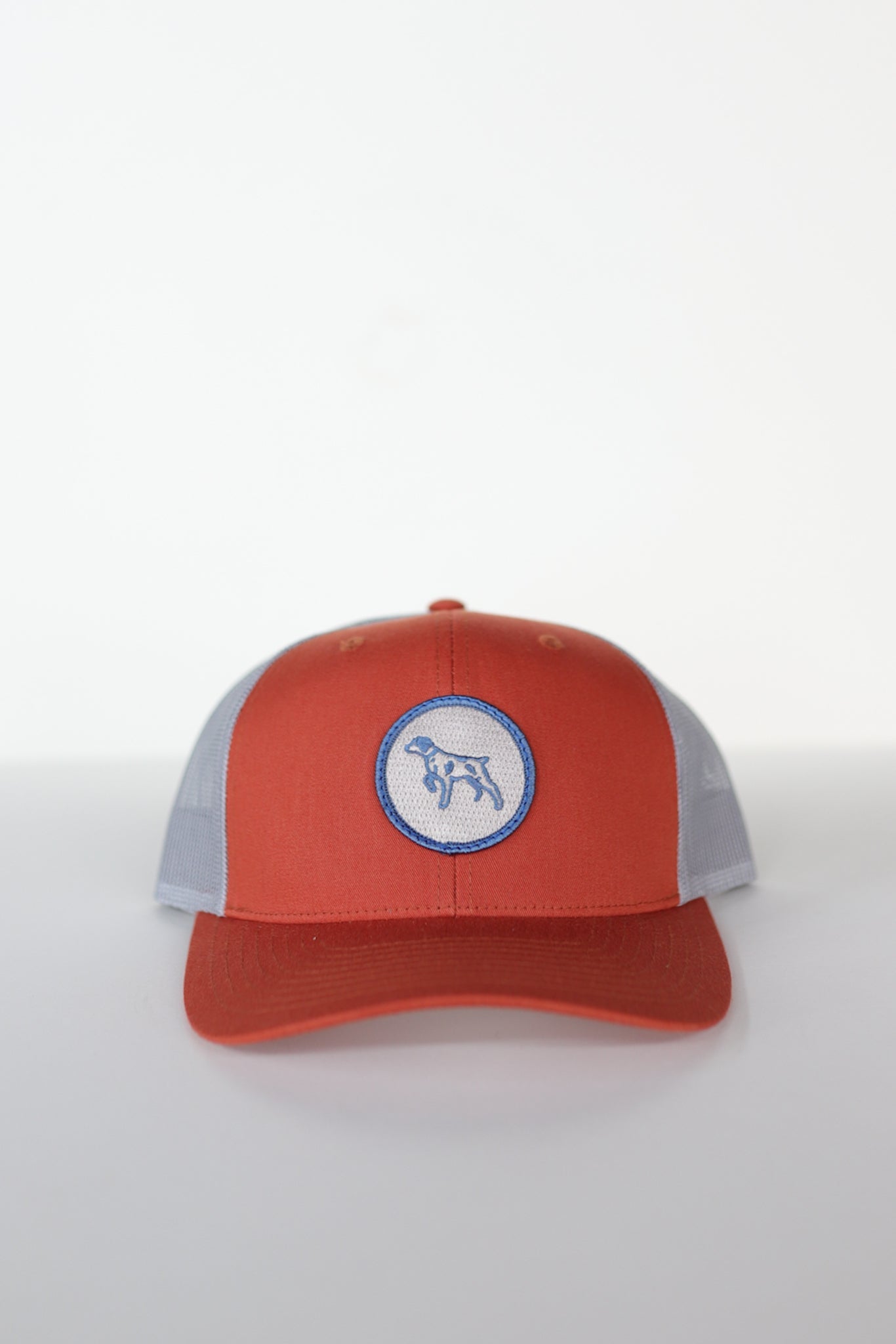 The Shop Hat | Merit Badge by Ballad of the Bird Dog is a red and gray snapback trucker hat featuring a circular patch with a bird dog silhouette on the front.