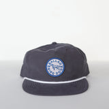 The Shop Hat | Merit Badge by Ballad of the Bird Dog is a dark snapback baseball cap featuring a blue and white circular patch with "BALLAD OF THE BIRD DOG" and a central dog graphic on the front.