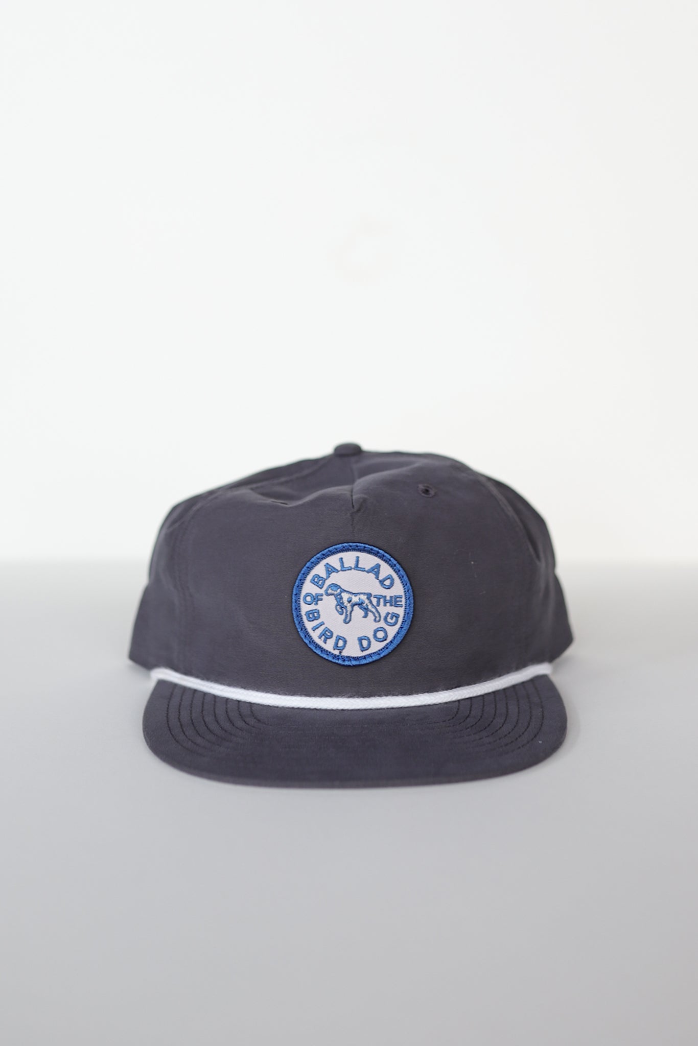 The Shop Hat | Merit Badge by Ballad of the Bird Dog is a dark snapback baseball cap featuring a blue and white circular patch with "BALLAD OF THE BIRD DOG" and a central dog graphic on the front.