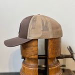 The Ballad of the Bird Dog Shop Hat | Merit Badge is a brown and tan mesh snapback trucker hat, shown on a wooden hat block with a metal clamp against a plain background.