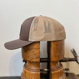 The Shop Hat | Merit Badge by Ballad of the Bird Dog, a brown and tan mesh snapback trucker hat, is shown sideways on a vintage wooden stretcher against a plain background, highlighting its merit badge patch.