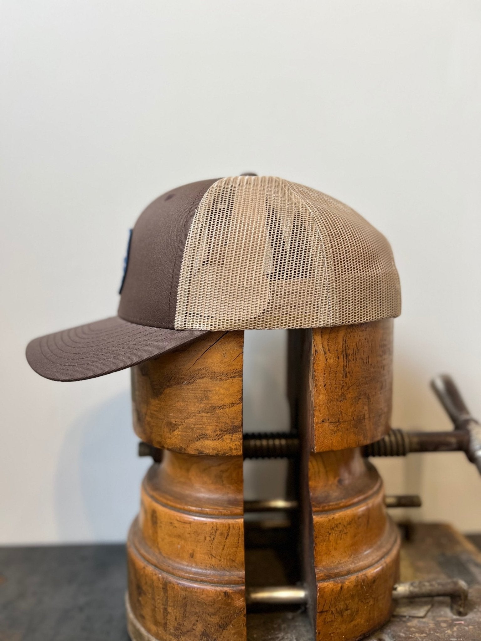 The Shop Hat | Merit Badge by Ballad of the Bird Dog, a brown and tan mesh snapback trucker hat, is shown sideways on a vintage wooden stretcher against a plain background, highlighting its merit badge patch.