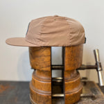 The Shop Hat | Merit Badge by Ballad of the Bird Dog, a light brown snapback cap, is held upright in a large wooden vise on a dark surface against a plain background.