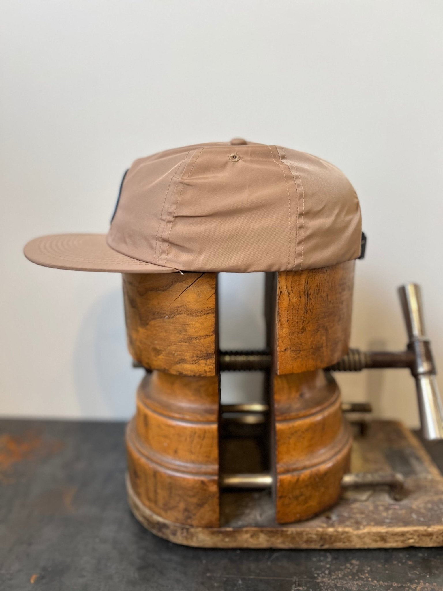 The Shop Hat | Merit Badge by Ballad of the Bird Dog is shown as a beige snapback cap on a large wooden clamp against a plain background.