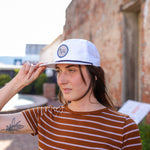 A person in a brown and white striped shirt stands outside near a brick wall, touching the brim of the Shop Hat | Merit Badge by Ballad of the Bird Dog.