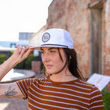 A woman outdoors in a brown striped shirt and white snapback touches the brim of her Ballad of the Bird Dog Shop Hat | Merit Badge while standing by a brick wall and looking off to the side.