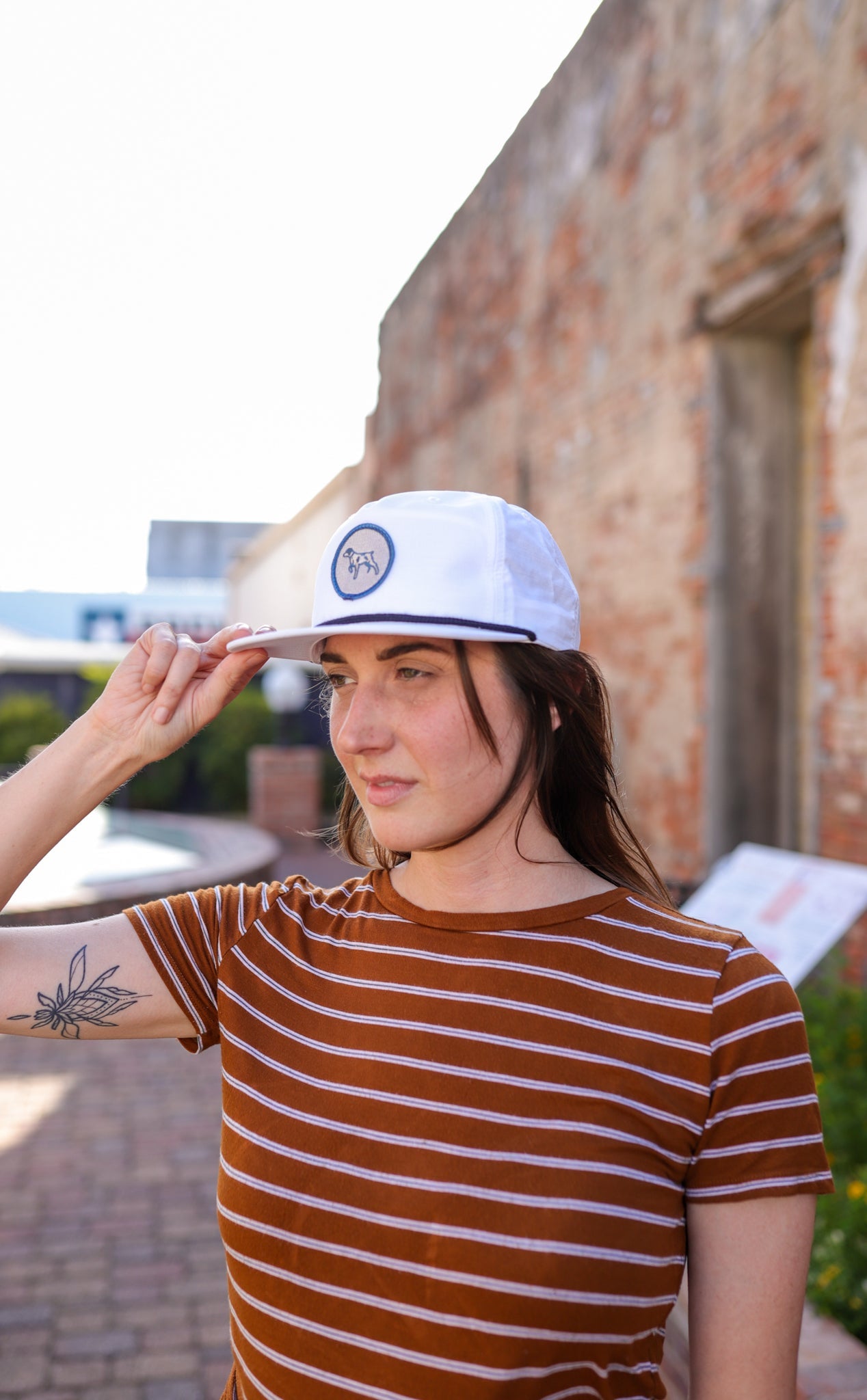 A woman outdoors in a brown striped shirt and white snapback touches the brim of her Ballad of the Bird Dog Shop Hat | Merit Badge while standing by a brick wall and looking off to the side.