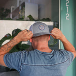 A man wearing the Ballad of the Bird Dog Shop Hat | Merit Badge snapback, in gray and white, faces away as he adjusts his hat near a door marked 212.