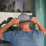 A person in a blue t-shirt wears the Ballad of the Bird Dog Shop Hat | Merit Badge, adjusting it while standing in front of a green door labeled 212. A merit badge patch is also visible on their sleeve.