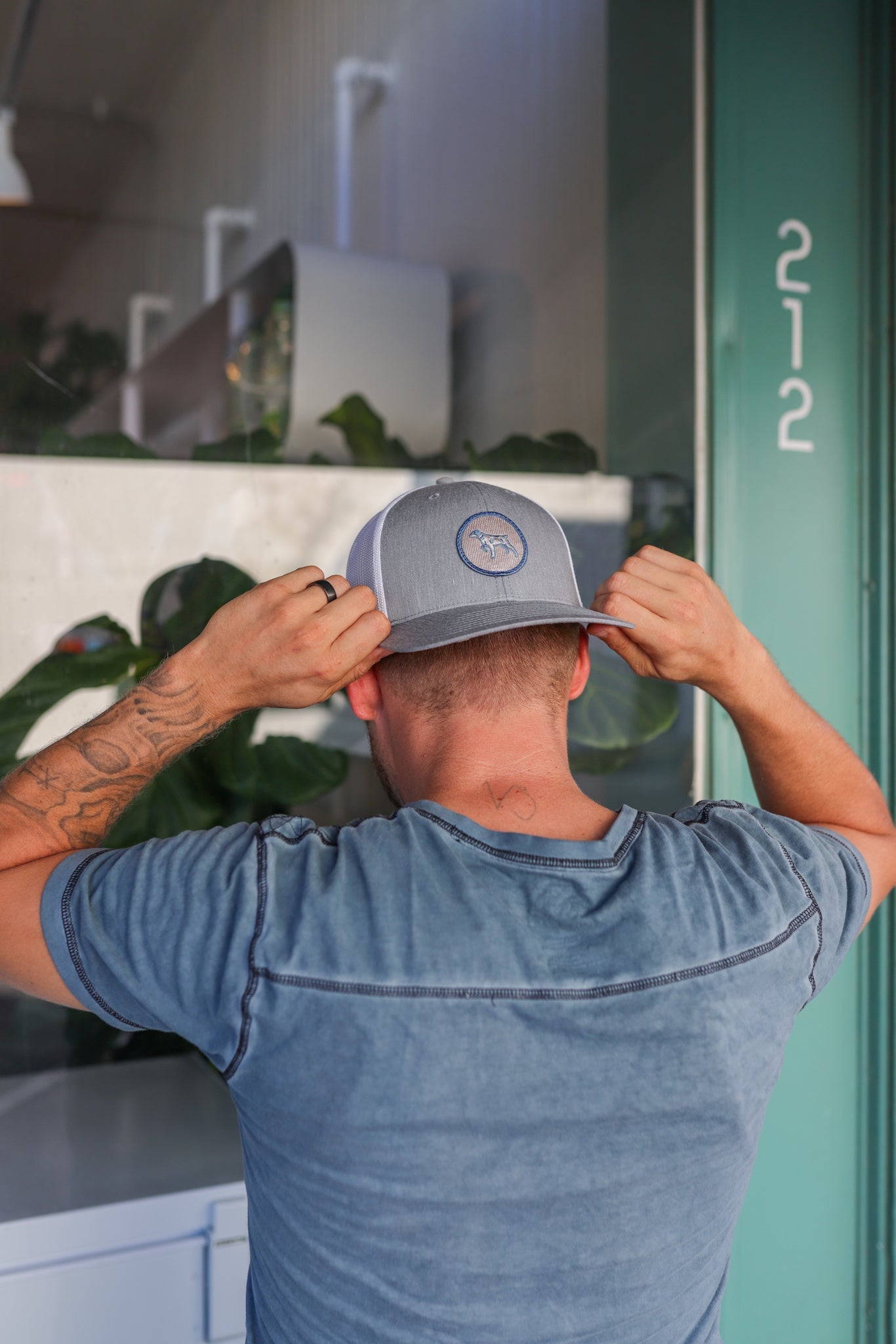 A person in a blue t-shirt wears the Ballad of the Bird Dog Shop Hat | Merit Badge, adjusting it while standing in front of a green door labeled 212. A merit badge patch is also visible on their sleeve.