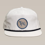 The Ballad of the Bird Dog Shop Hat | Merit Badge is a white snapback cap featuring a blue bird dog outline badge on the front and a slim dark line above the brim.