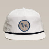 The Shop Hat | Merit Badge by Ballad of the Bird Dog is a white baseball cap with black brim trim and a blue-embroidered circular bird dog patch on the front.
