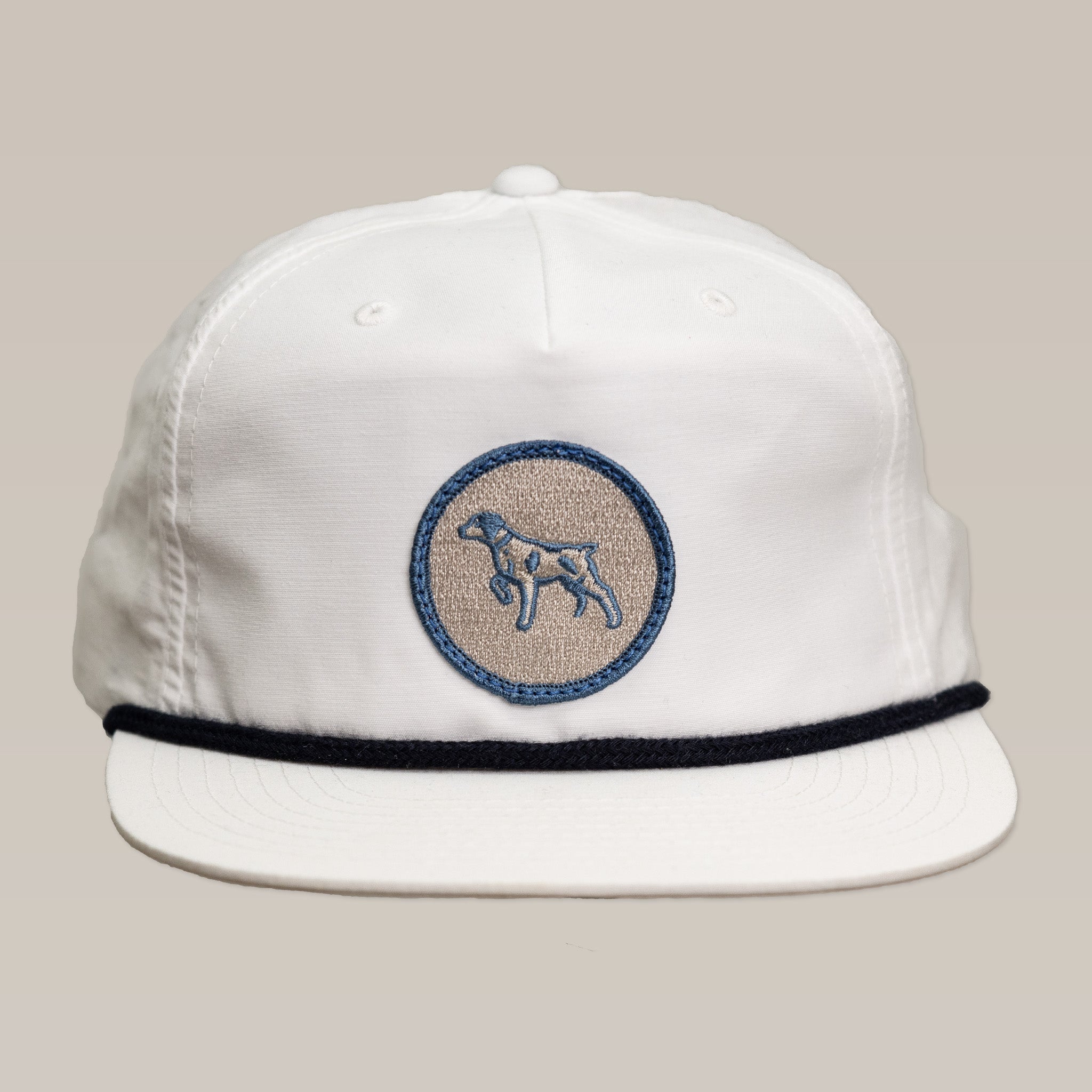The Shop Hat | Merit Badge by Ballad of the Bird Dog is a white baseball cap with black brim trim and a blue-embroidered circular bird dog patch on the front.