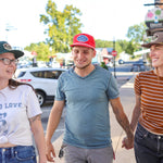 Three people wearing casual clothes and Shop Hat | Merit Badge by Ballad of the Bird Dog walk together on a sidewalk, smiling, with cars and trees in the background.