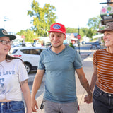 Three friends wear hats from Ballad of the Bird Dog, including the Shop Hat | Merit Badge, as they walk together on a sunny sidewalk. The man in the middle smiles while the two women beside him hold hands and look at each other.