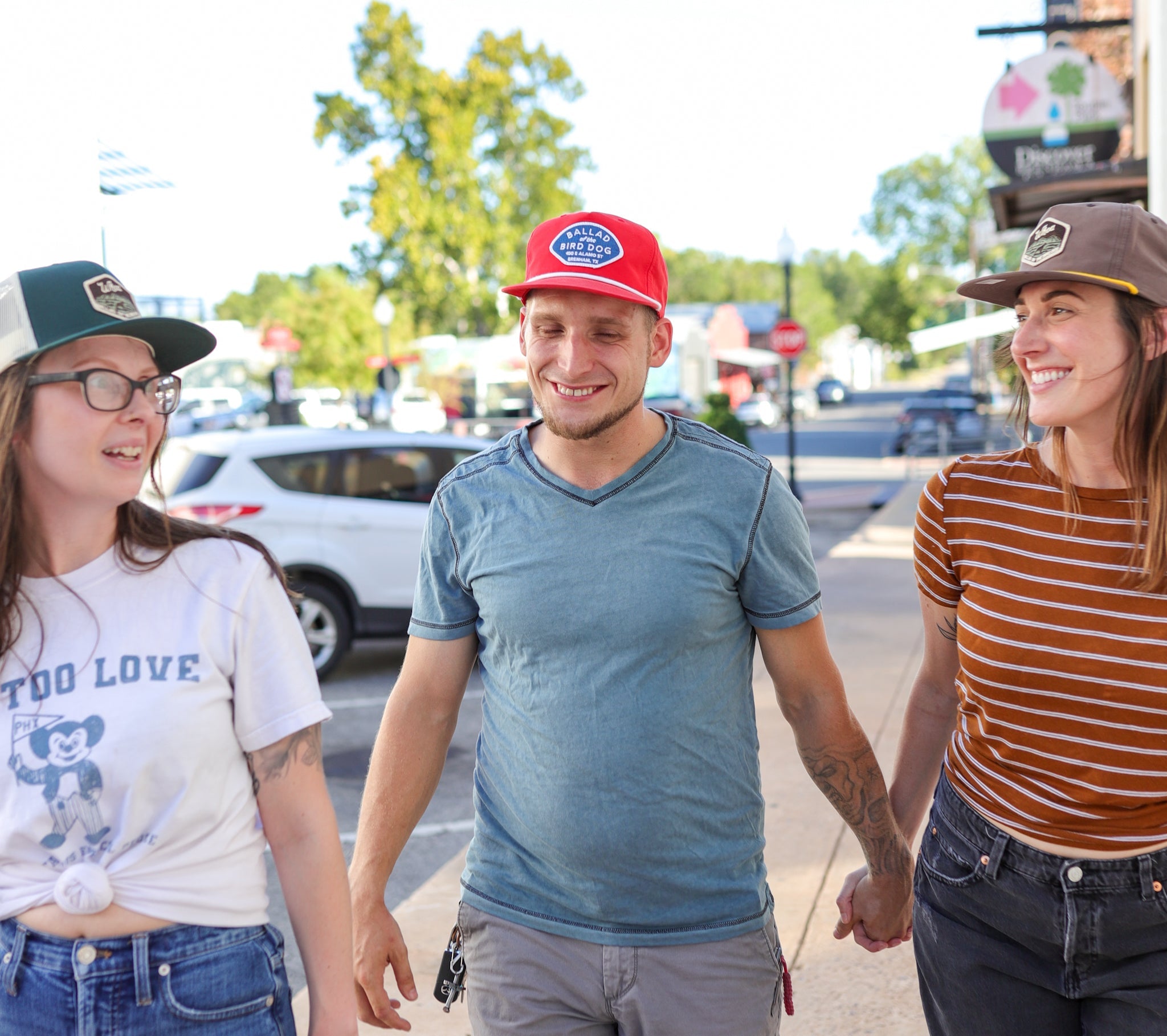 Three friends wear hats from Ballad of the Bird Dog, including the Shop Hat | Merit Badge, as they walk together on a sunny sidewalk. The man in the middle smiles while the two women beside him hold hands and look at each other.