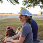 A woman wearing the Shop Hat | Merit Badge by Ballad of the Bird Dog sits on a wooden bench outdoors, smiling with two people beside her. She pairs it with a gray shirt and pink shorts; trees and a white fence are in the background.