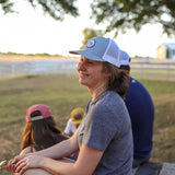 A woman in a gray shirt and pink shorts wears the Ballad of the Bird Dog "Shop Hat | Merit Badge" snapback while sitting outdoors on a wooden platform, smiling with friends. Trees and a fence can be seen in the background.