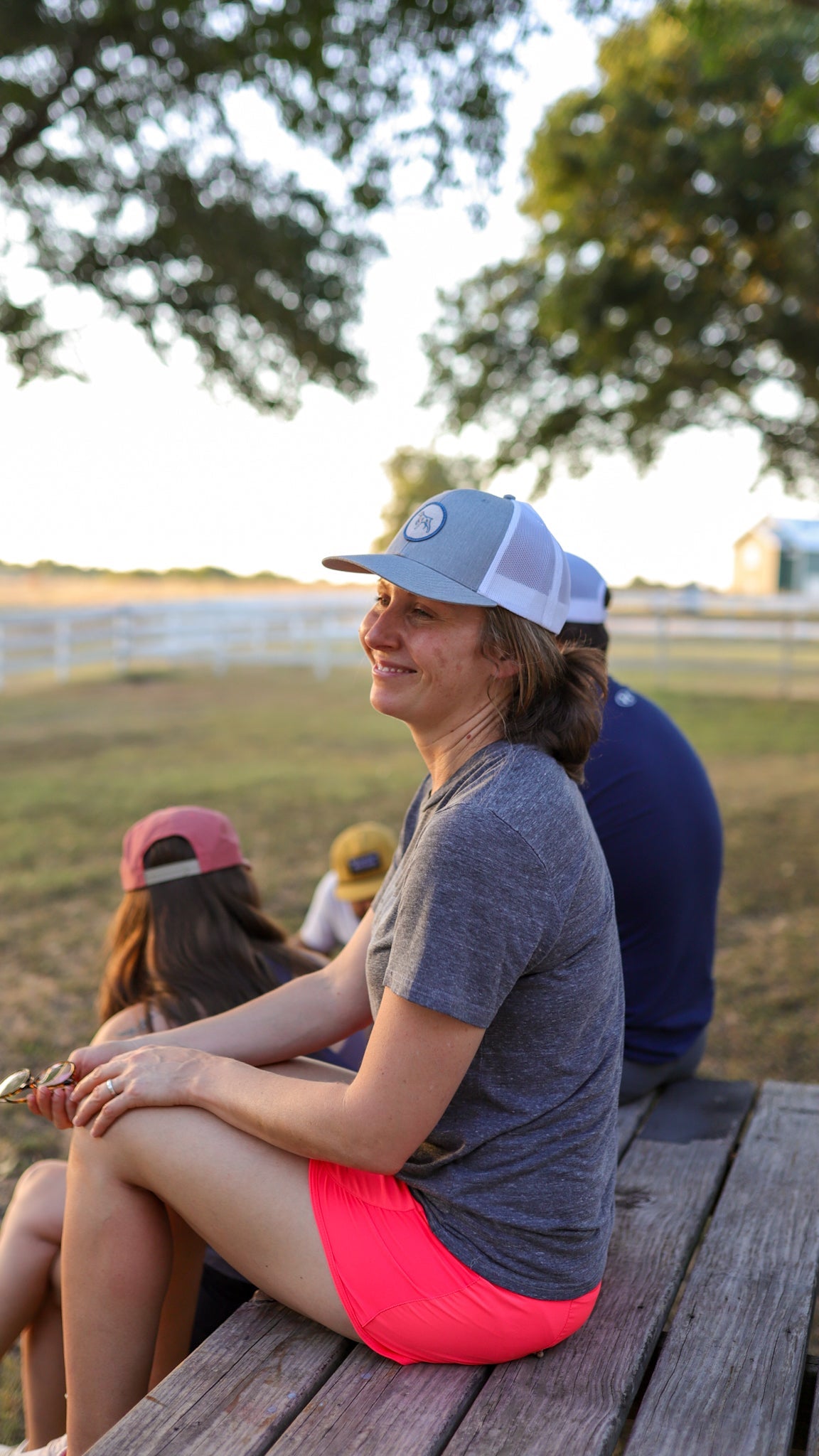 A woman in a gray shirt and pink shorts wears the Ballad of the Bird Dog "Shop Hat | Merit Badge" snapback while sitting outdoors on a wooden platform, smiling with friends. Trees and a fence can be seen in the background.