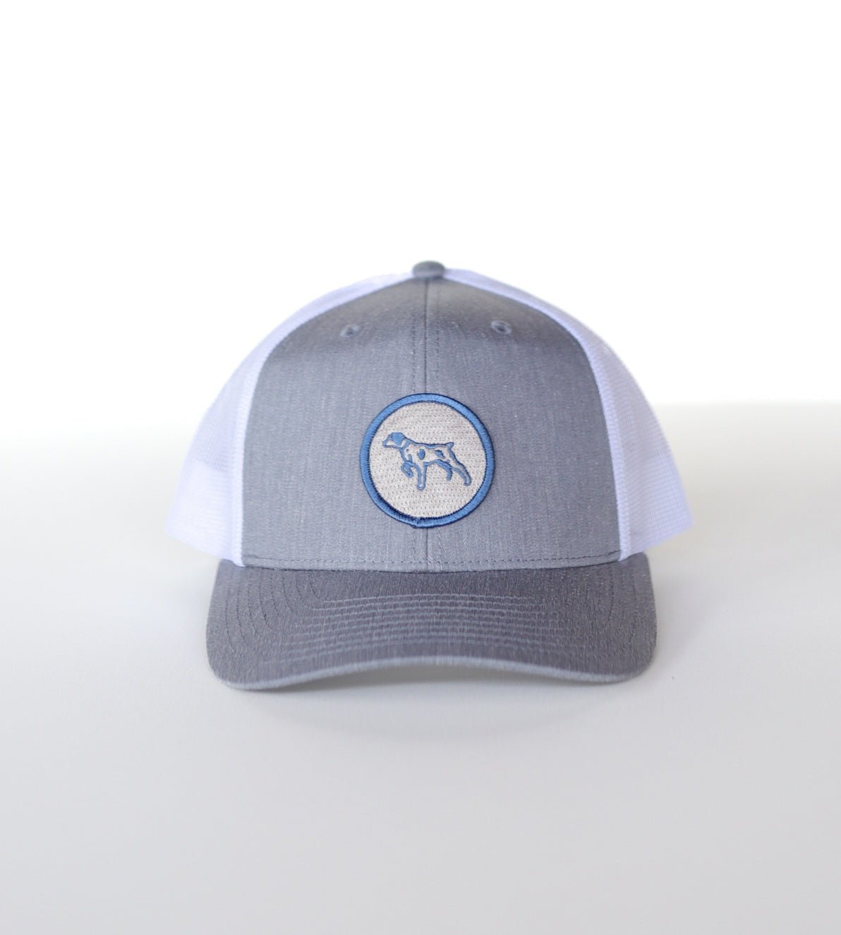 The Shop Hat | Merit Badge by Ballad of the Bird Dog is a grey and white snapback trucker hat featuring a circular merit badge patch with a bird dog outline on the front panel.