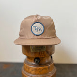 The Ballad of the Bird Dog Shop Hat | Merit Badge is a tan snapback baseball cap with a circular patch featuring a running bird dog, displayed on a wooden hat stand against a plain background.