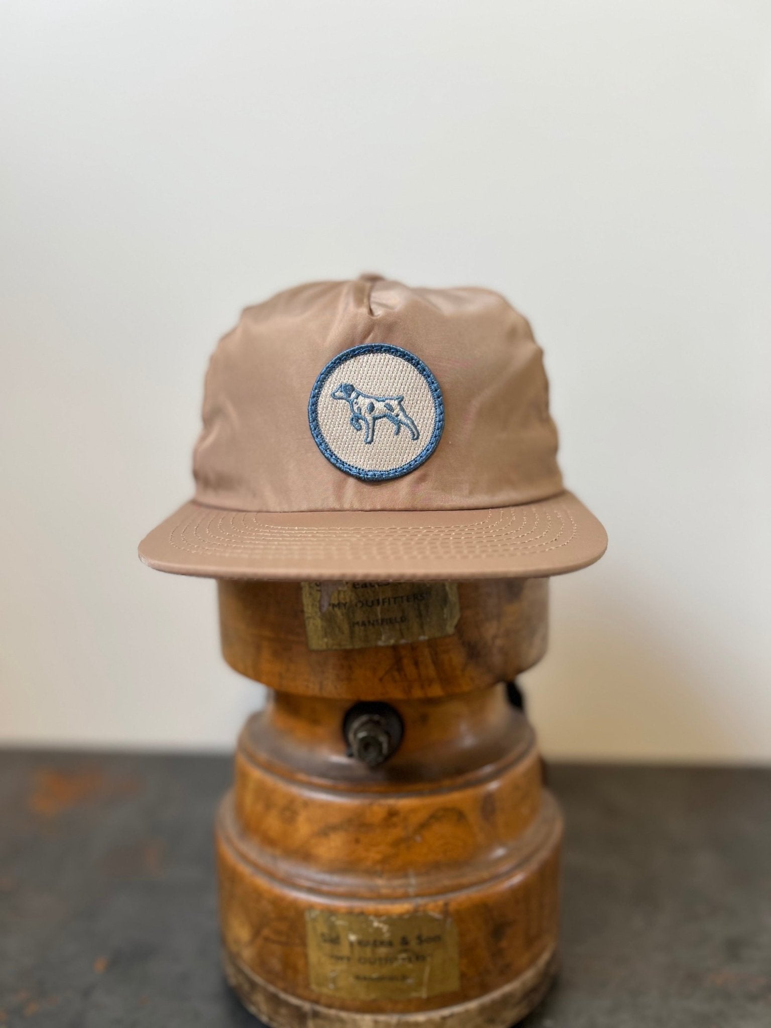 The Ballad of the Bird Dog Shop Hat | Merit Badge is a tan snapback baseball cap with a circular patch featuring a running bird dog, displayed on a wooden hat stand against a plain background.