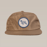 The Ballad of the Bird Dog Shop Hat | Merit Badge is a brown snapback cap with a round patch featuring a blue outlined bird dog on the front, shown on a beige background.