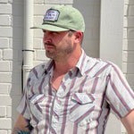 A man wearing a light green Shop Hat | Neon Revival by Ballad of the Bird Dog and a striped short-sleeve shirt stands against a white brick wall in sunlight.