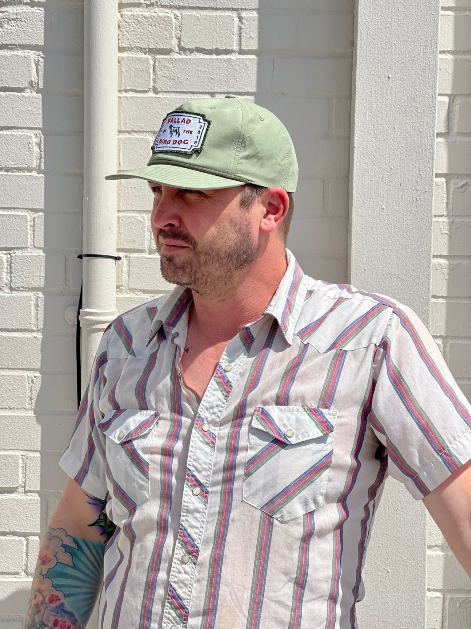 A man in a striped short-sleeve shirt and the Ballad of the Bird Dog Shop Hat | Neon Revival stands against a white brick wall in downtown Brenham, sunlight highlighting his arm tattoo.