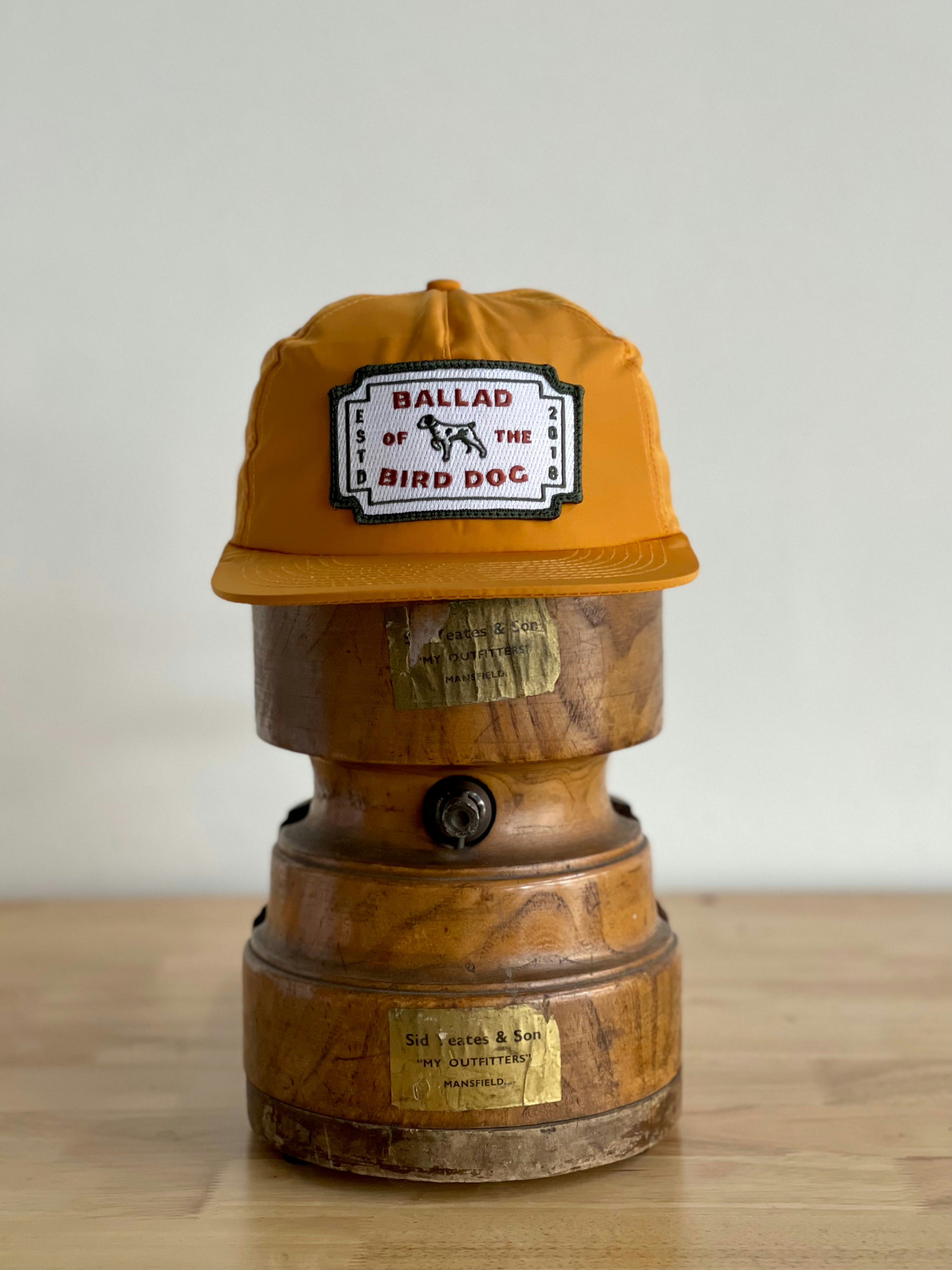 The Shop Hat | Neon Revival by Ballad of the Bird Dog, featuring the classic logo patch, sits on a vintage wooden object with brass labels—reflecting downtown Brenham style.