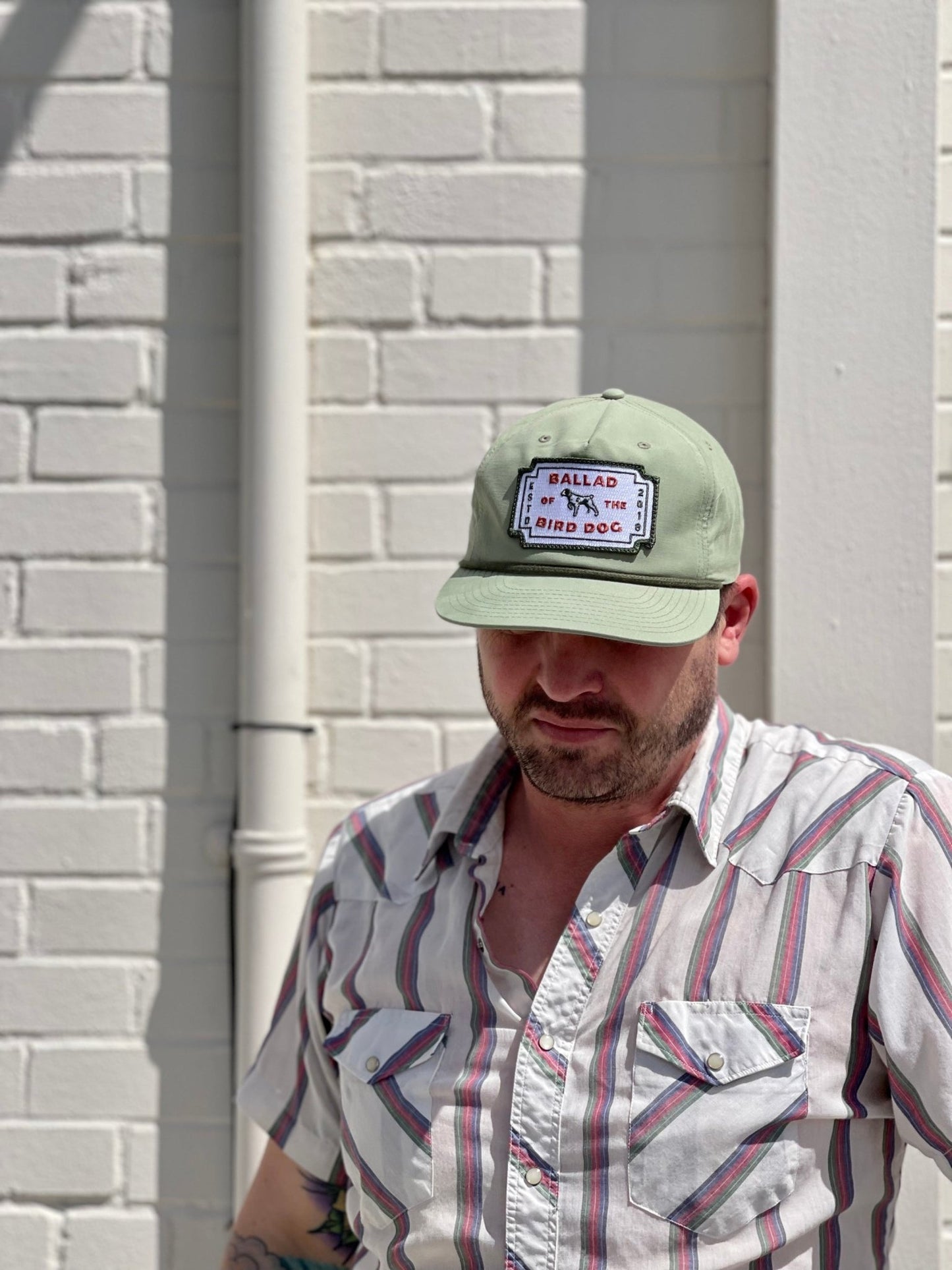 A man stands against a white brick wall in sunlight, wearing a striped short-sleeve shirt and the Ballad of the Bird Dog Shop Hat | Neon Revival—a classic cap showing off downtown Brenham style with a signature patch.