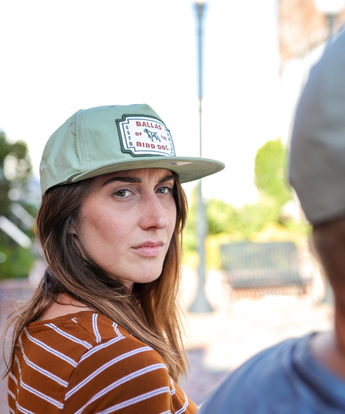 A woman in a striped shirt and a Ballad of the Bird Dog Shop Hat | Neon Revival looks over her shoulder outdoors in downtown Brenham; another person is partially visible in the foreground.