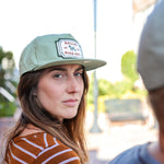 A woman in a striped shirt and a Ballad of the Bird Dog Neon Revival Shop Hat looks over her shoulder outdoors, while another person appears blurred in the foreground.