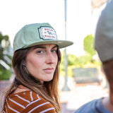 A woman in a striped shirt and a Ballad of the Bird Dog Shop Hat | Neon Revival looks over her shoulder outdoors in downtown Brenham; another person is partially visible in the foreground.