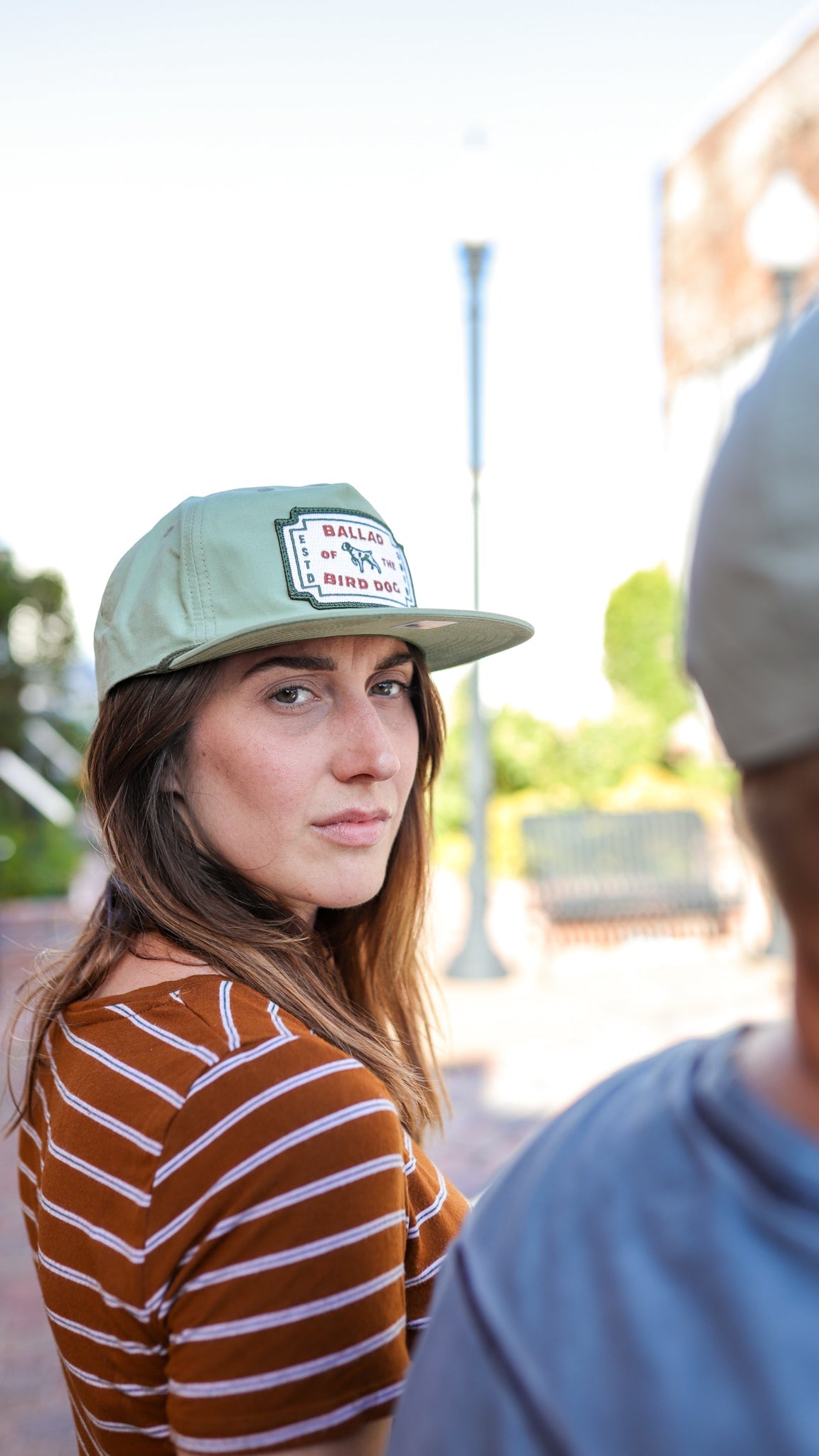 A woman in a striped shirt and a Ballad of the Bird Dog Shop Hat | Neon Revival looks over her shoulder outdoors in downtown Brenham; another person is partially visible in the foreground.