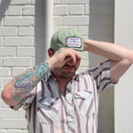 A person in a striped short-sleeve shirt and green Shop Hat | Neon Revival by Ballad of the Bird Dog stands against a white brick wall, shading their eyes with one hand. Their left arm features a colorful tattoo.