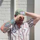 A person with a colorful tattoo adjusts their Ballad of the Bird Dog Shop Hat | Neon Revival, standing against a white brick wall in a striped short-sleeve shirt, reflecting the lively spirit of downtown Brenham.