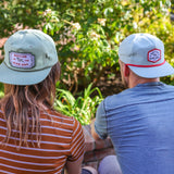 Two people wearing Ballad of the Bird Dog Shop Hat | Neon Revival sit side by side on a brick wall in downtown Brenham, facing away and enjoying a laid-back moment, with sunlight and greenery in the background.