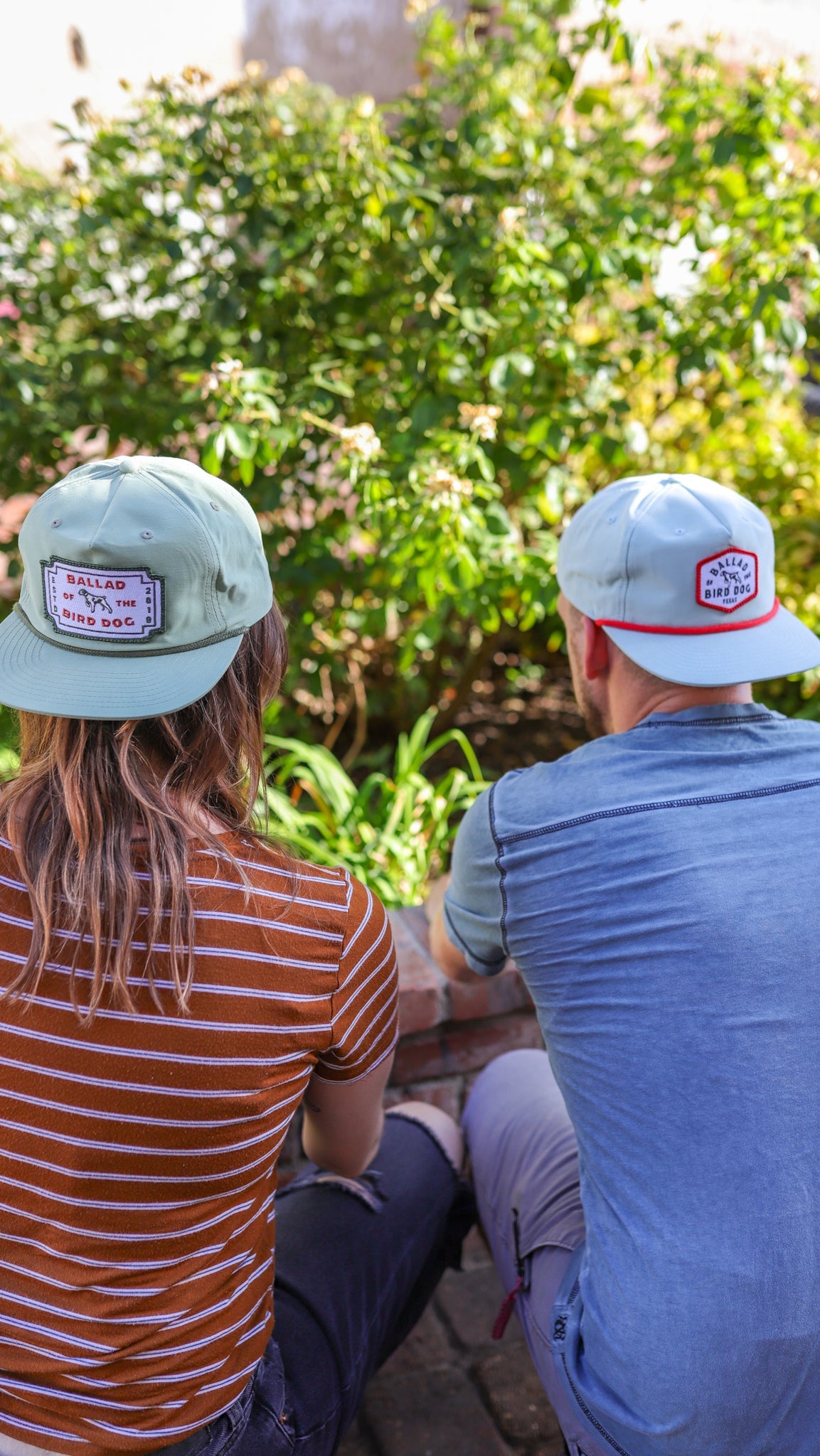 Two people wearing Ballad of the Bird Dog Shop Hat | Neon Revival sit side by side on a brick wall in downtown Brenham, facing away and enjoying a laid-back moment, with sunlight and greenery in the background.