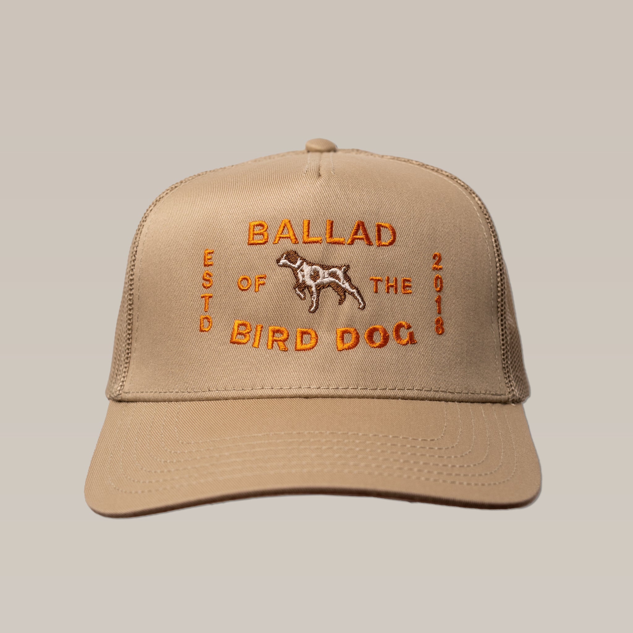 The Shop Hat | Neon Sign by Ballad of the Bird Dog is a tan trucker hat with orange "BALLAD OF THE BIRD DOG," "ESTD 2018," and a dog outline embroidered on the front.