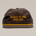 The Shop Hat | Old Fashioned by Ballad of the Bird Dog is a brown western hat with yellow trim and "BALLAD OF THE BIRD DOG" embroidered on the front—a perfect rustic accessory for casual outfits.