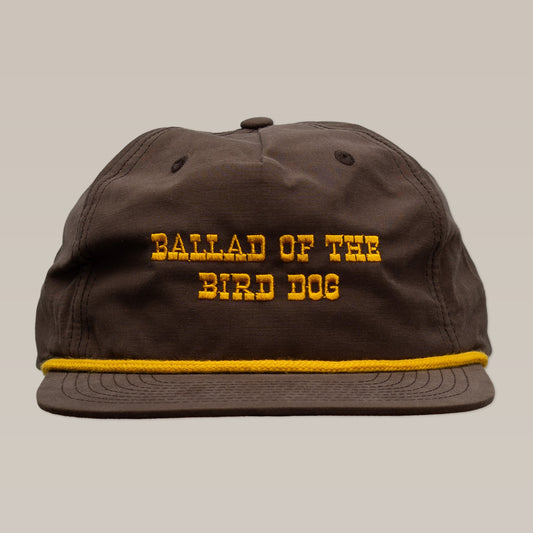 The Shop Hat | Old Fashioned by Ballad of the Bird Dog is a brown hat featuring yellow "BALLAD OF THE BIRD DOG" embroidery and a yellow rope above the brim—a classic Wild West-inspired accessory.