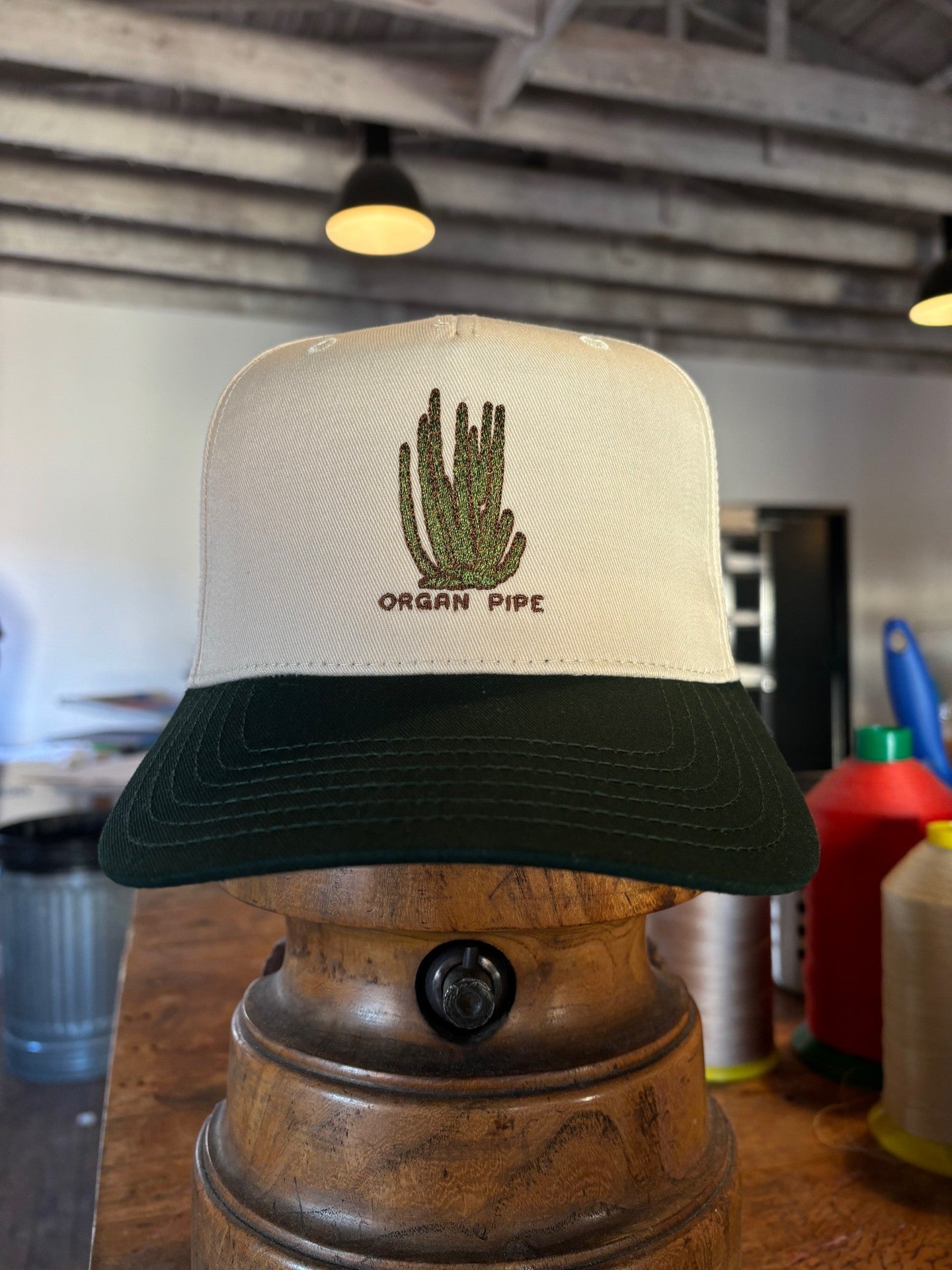 The Shop Hat | Organ Pipe by Ballad of the Bird Dog is a white and green trucker hat with an embroidered cactus and the words "Organ Pipe," shown on a wooden stand indoors.