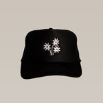 The Shop Hat | Peace Flower by Ballad of the Bird Dog is a black hat with three white embroidered peace flowers on the front, making it a stylish accessory that stands out beautifully against a plain beige background.