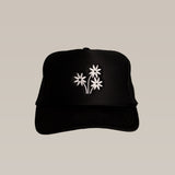 The Shop Hat | Peace Flower by Ballad of the Bird Dog is a black hat with three white embroidered peace flowers on the front, making it a stylish accessory that stands out beautifully against a plain beige background.