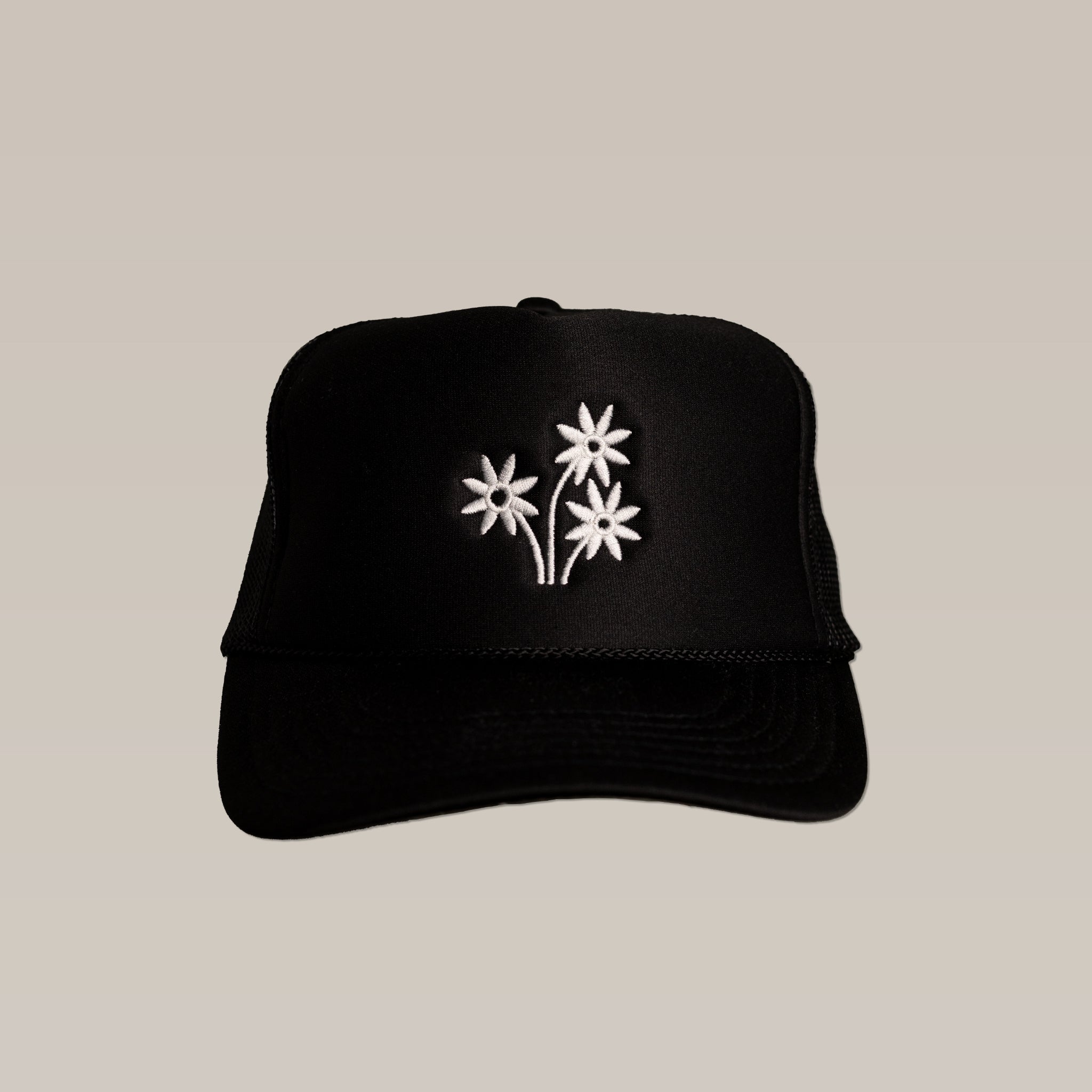 The Shop Hat | Peace Flower by Ballad of the Bird Dog is a black hat with three white embroidered peace flowers on the front, making it a stylish accessory that stands out beautifully against a plain beige background.