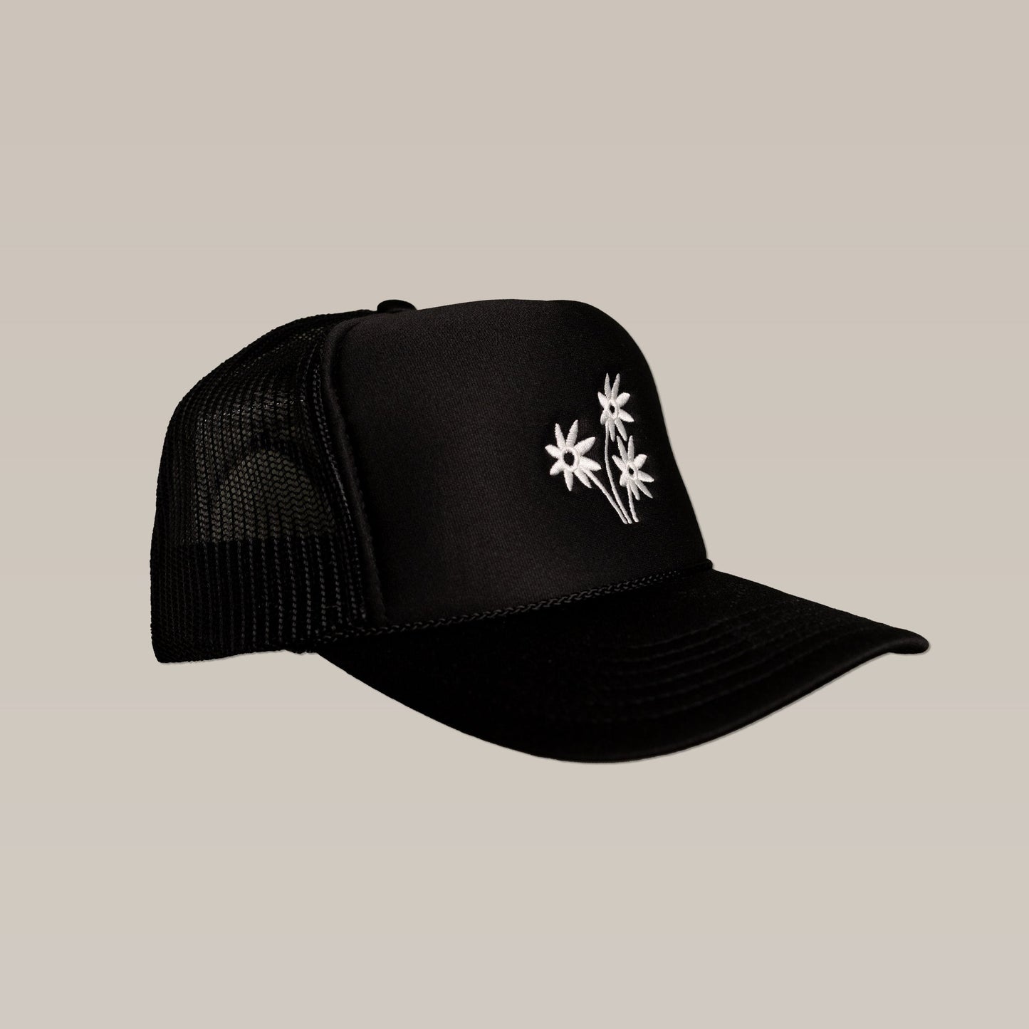The Shop Hat | Peace Flower by Ballad of The Bird Dog is a black mesh trucker cap with three white embroidered peace flowers on the front panel, shown on a plain beige background.